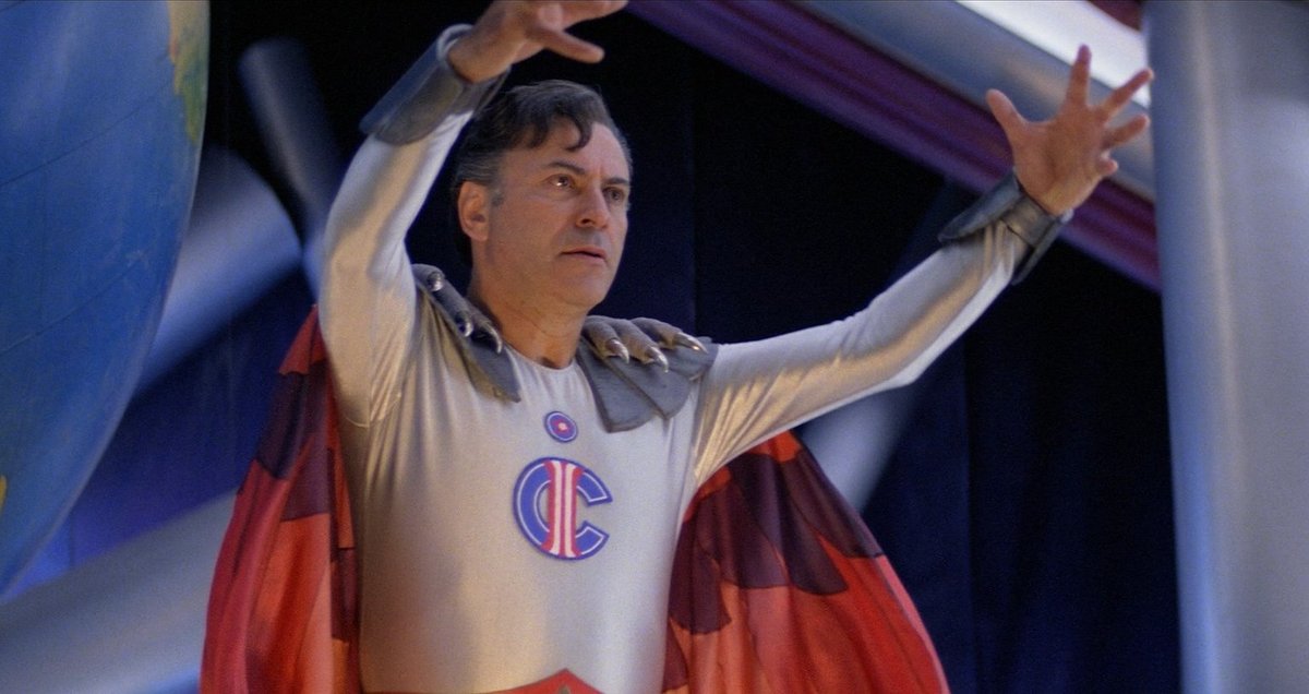 Alan Arkin wearing a goofy superhero costume with a logo that combines an I and a C in The Return of Captain Invincible.